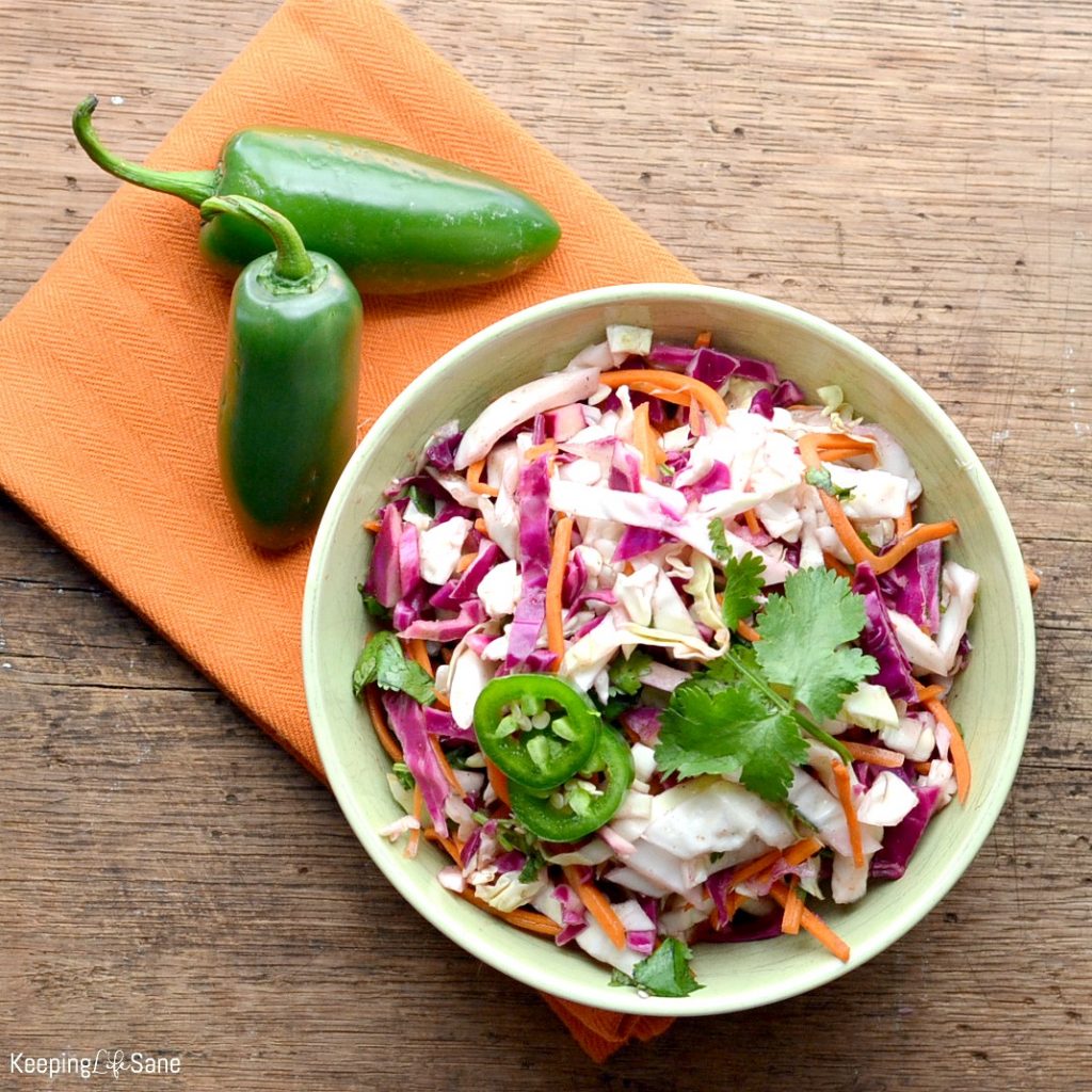Overhead view of white bowl filled with Mexican coleslaw with orange napkin and jalapenos.