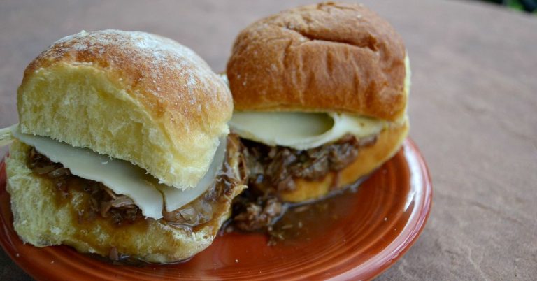 two slider roast beef sandwiches on dark red plate with meat adnd cheese and gravy dripping out.