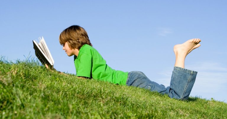 boy laying on grass in jeans and green shirt reading a book