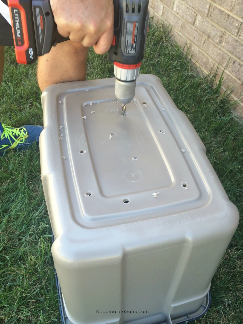 Plastic Rubbermaid bin that's upside down in grass with a man's hand drilling holes in the bottom.