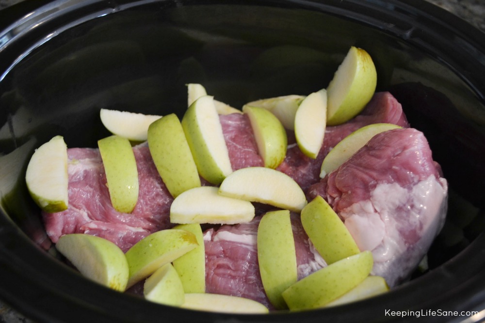 raw pork in a black slow cooker pot with sliced granny smith apples laying on top