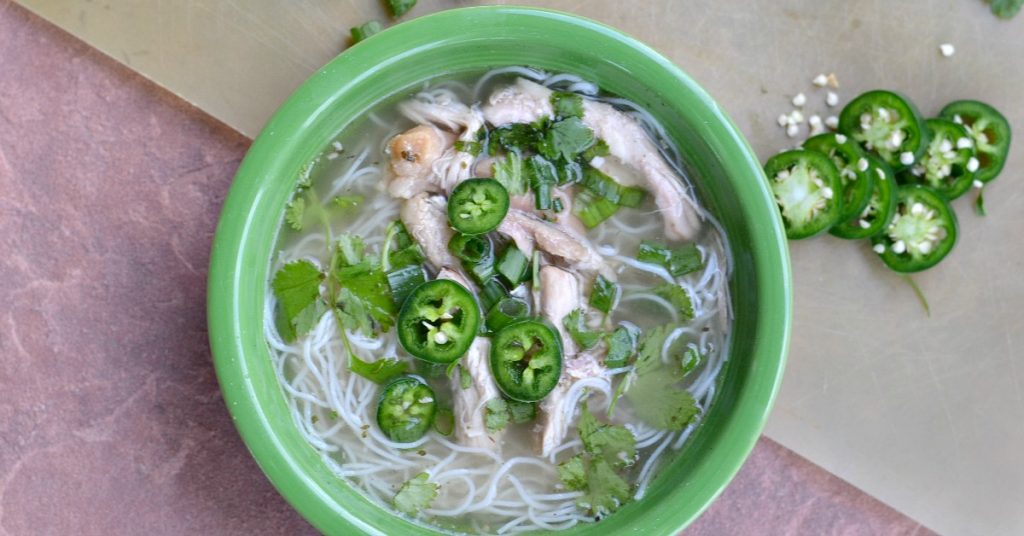 overhead view of homemade chicken pho in green bowl with noodles, chicken, cilantro, and jalapeños