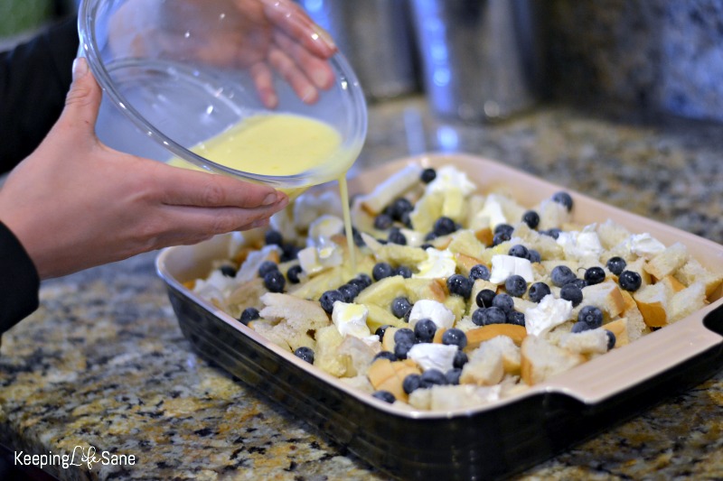 eggs being poured in casserole dish with bread, cream cheese, and blueberries