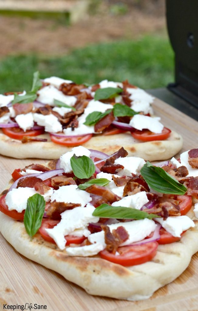 two pizzas on cutting board outside by grill with tomatoes, cheese, bacon and basil