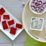 square cut red cranberry shots on white tray, round tray with red and green grapes, sugared cranberries in white bowl and blue cheese in square white bowl