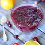 clear bowl of cranberry sauce from scratch on a wooden table with a spoon, fresh cranberries and oranges slices around it
