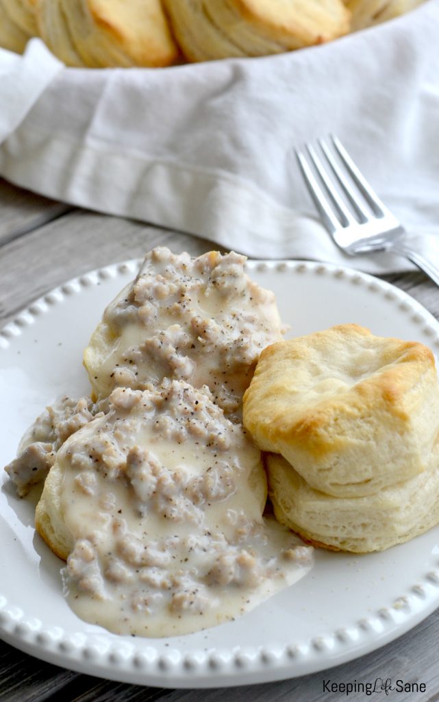 White plate with Southern biscuits and gravy on white plate with basket of cooked biscuits on wooden table