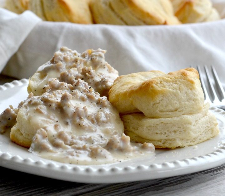 White plate with Southern biscuits and gravy on white plate with basket of cooked biscuits on wooden table
