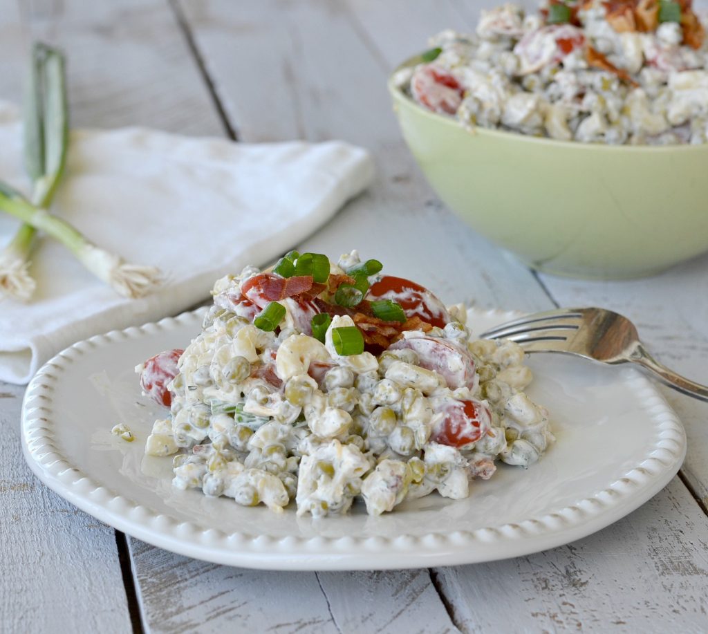 Creamy pea salad on white plate topped with crumbled bacon and chives with green bowl with pea salad in background