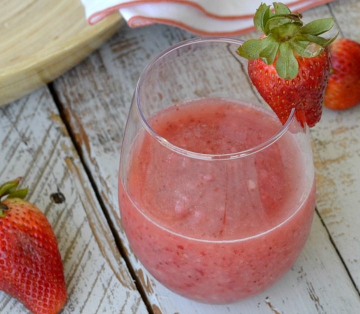 wine glass with frozen strawberry wine slushie on wooden boards with fresh strawberries laying about and one on the wine glass