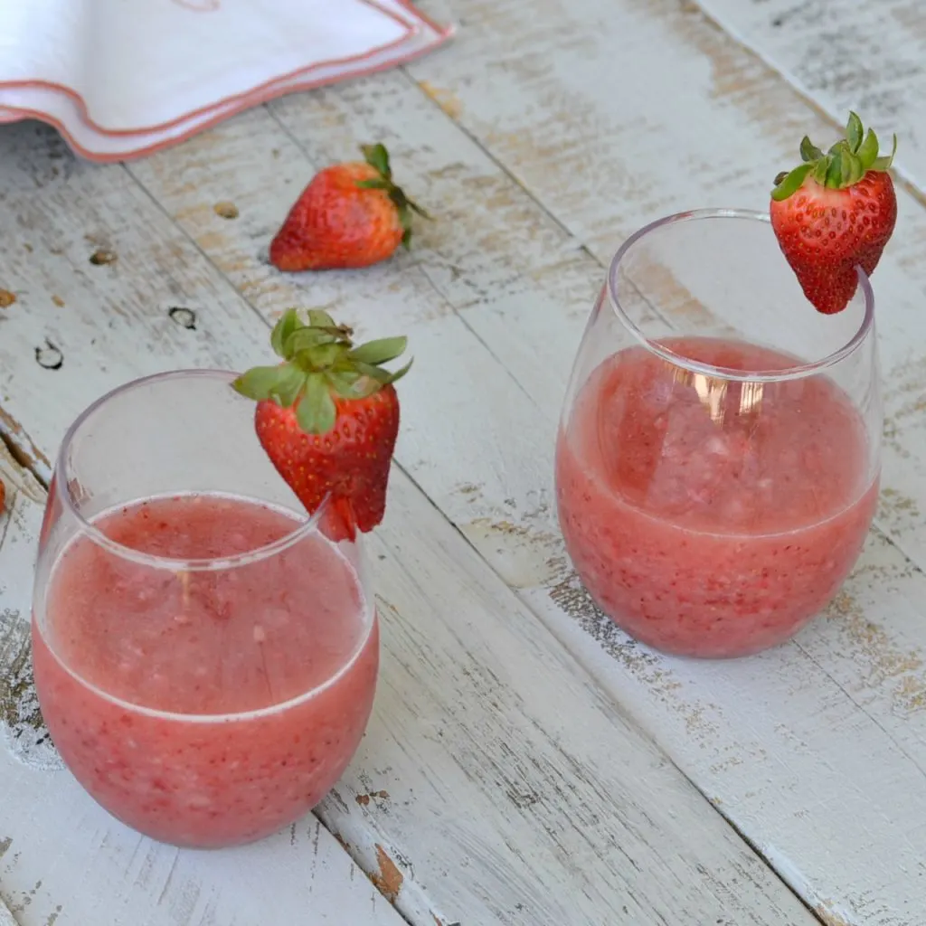 2 wine glasses with frozen strawberry wine slushie on wooden boards with fresh strawberries laying about and one on the wine glass
