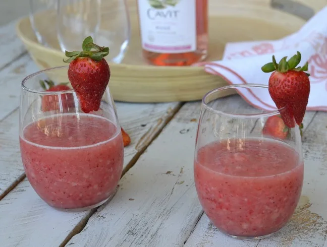 2 wine glasses with frozen strawberry wine slushie on wooden boards with fresh strawberries laying about and one on the wine glasses with tray with bottle of rose in the background