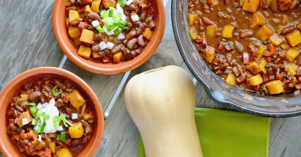 Cast iron pot with butternut squash chili and 2 bowls of chili topped with sour cream and green onions