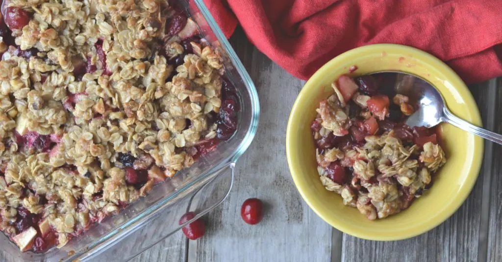 overhead view of apple cranberry crisp in yellow bowl on wooden table with baking pan