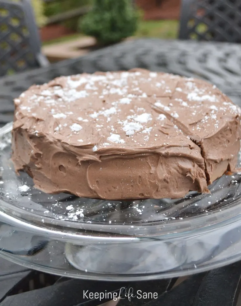 Round chocolate cake on glass cake stand with powder sugars sprinkled on top.