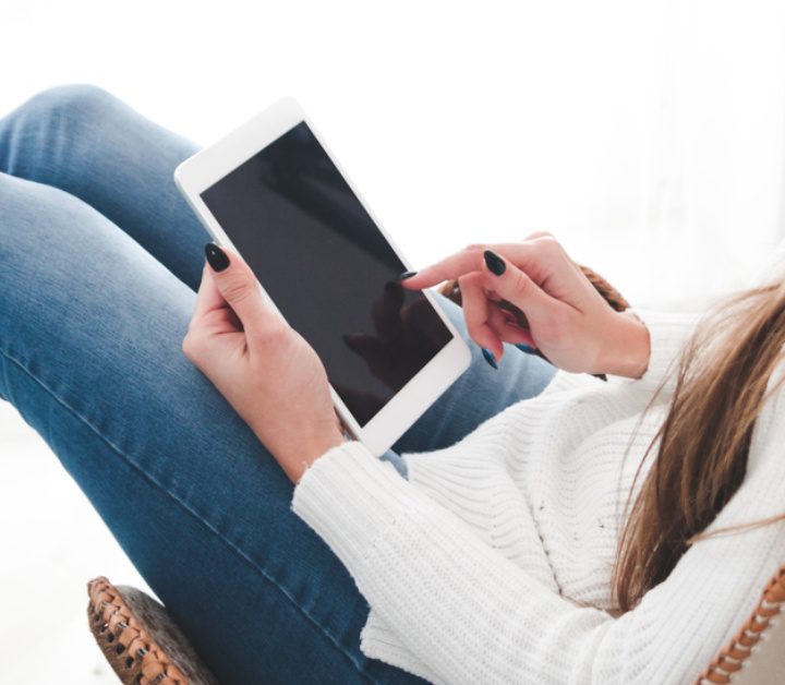 lady in jeans and white sweater sitting in chair and reading on a tablet