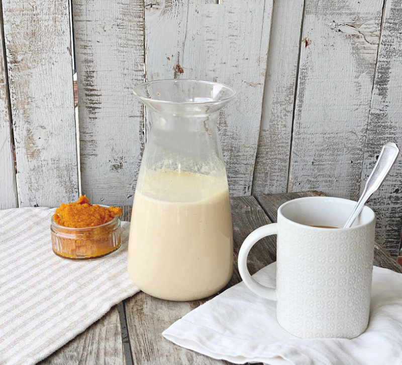 bottle with pumpkin spice creamer half full in front of grey wood background.  Cup of coffee with spoon and small bowl of pumpkin puree.