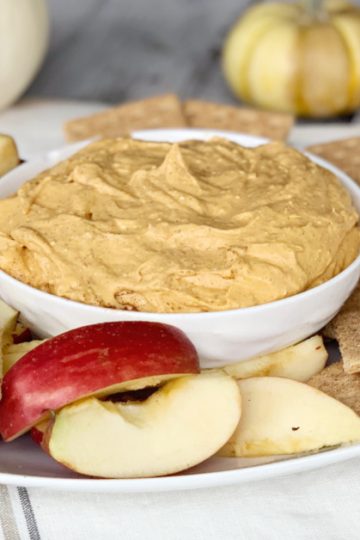 pumpkin pie dip with cream cheese in white bowl on fall tablecloth with apple slice and graham crackers