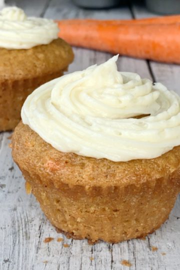2 eggless carrot cake cupcakes on wooden tables with 2 orange carrots in the background