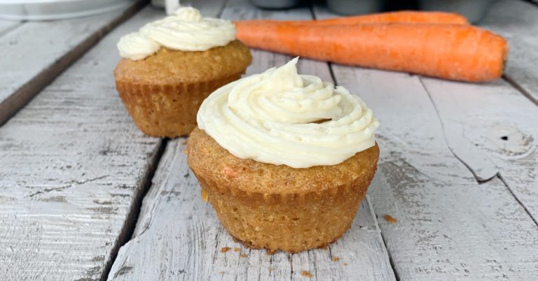 2 eggless carrot cake cupcakes on wooden tables with 2 orange carrots in the background
