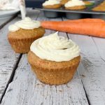 2 eggless carrot cake cupcakes on wooden tables with 2 orange carrots in the background