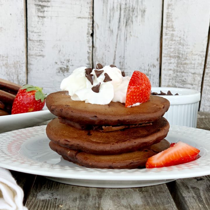 a stack of eggless chocolate pancakes on a white plate with whipped cream and chocolate chips and strawberries