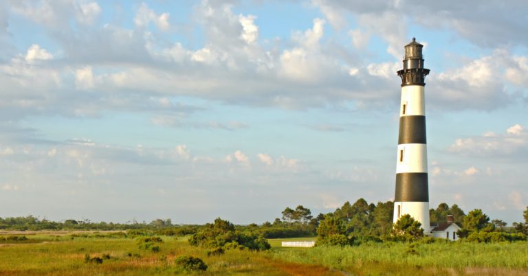 Walkway leading to the Bodie Island lighthouse over marshes of the Cape Hatteras National Seashore against white clouds and a blue morning sky