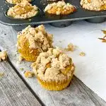 2 pumpkin muffins with crumbles on top with muffin pan in background