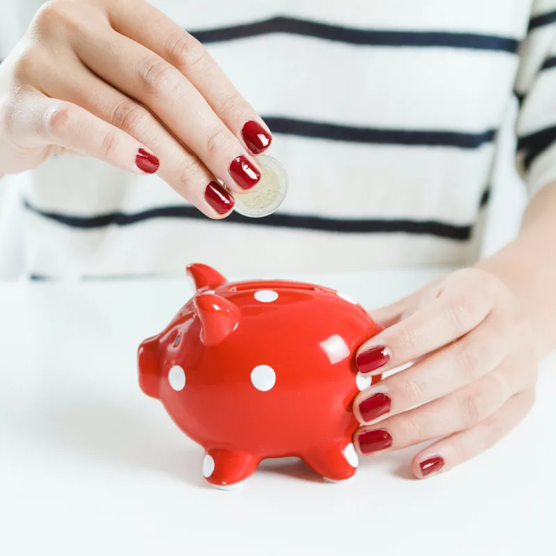 Lady with red fingernails adding a coin to a small red piggy bank.