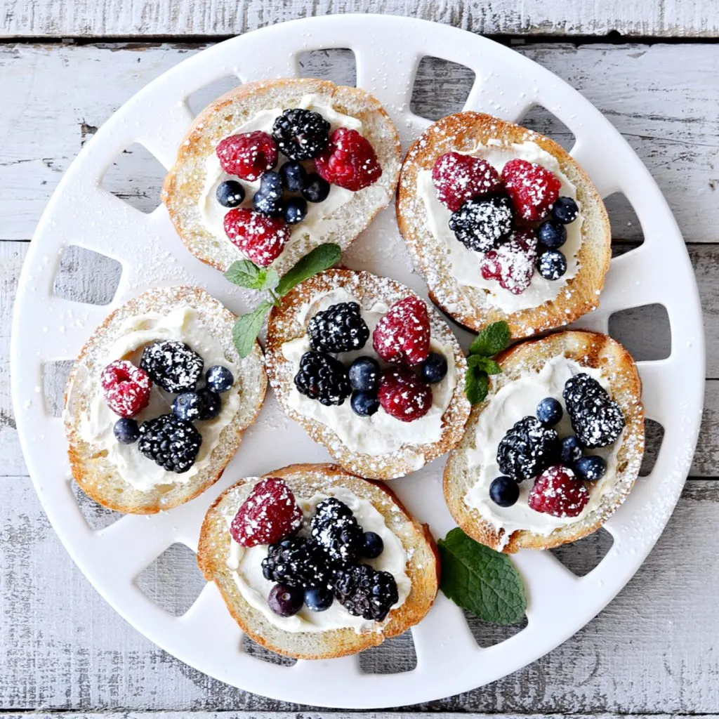 overhead view of berry bruschetta on round white platter- toast french bread with cream cheese spread with blueberries, blackberries and raspberries