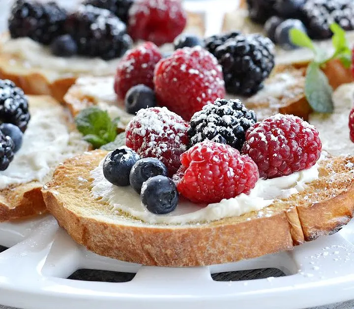 closeup view of berry bruschetta on round white platter- toast french bread with cream cheese spread with blueberries, blackberries and raspberries