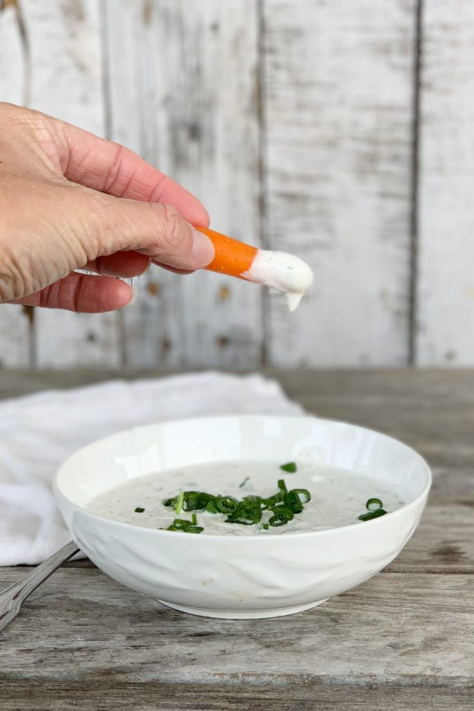 ranch dip in white bowl with scallions on top with hand holding baby carrot that's been dipped