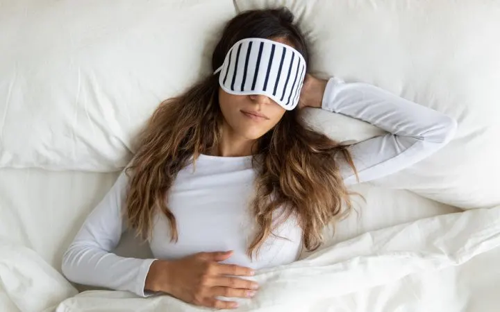 lady with long brown hair sleeping on white bed in white long sleeved tee with black and white eye mask