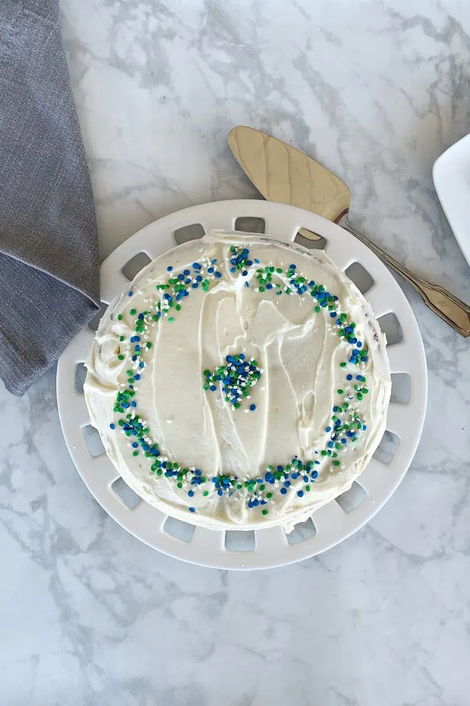 overhead view of vanilla cake on maple countertop on round cake plate. Cake has blue, green and white sprinkles