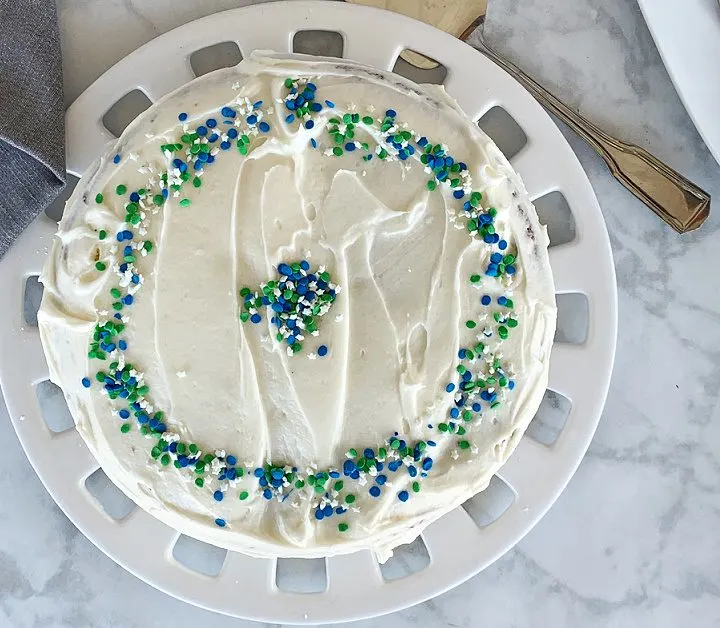 overhead view of vanilla cake on maple countertop on round cake plate. Cake has blue, green and white sprinkles