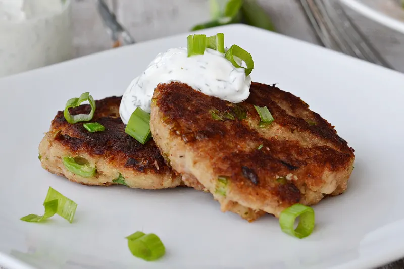 closeup of two tuna patties on a white plate with sour cream dill topping and chopped chives