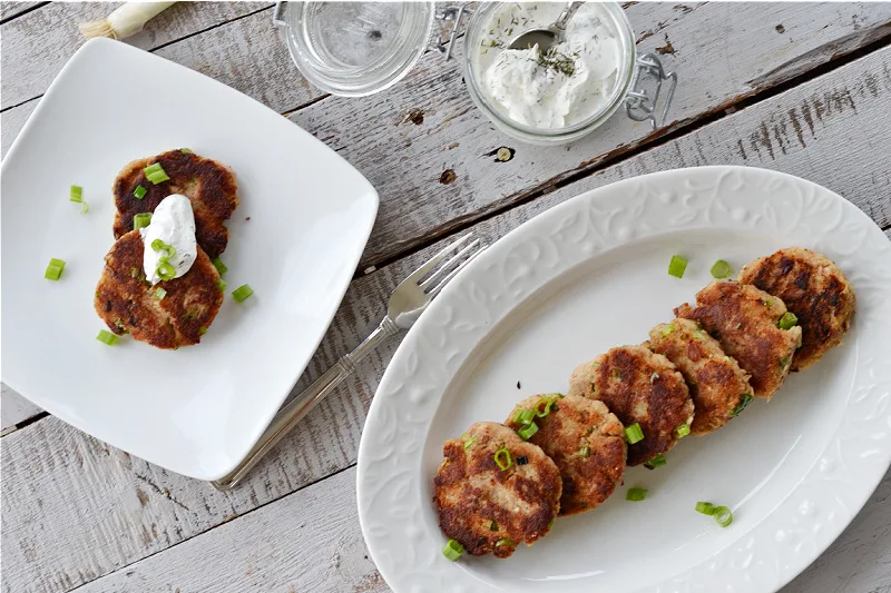 overhead view of two tuna patties on a white plate with sour cream dill topping and chopped chives and white platter with 6 patties on it