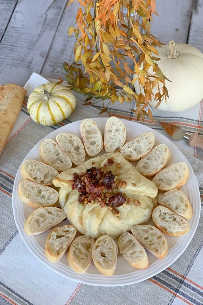 overhead view baked brie with maple syrup in puff pastry on white plate with crispy bacon and walnuts on top and sliced bread around it.