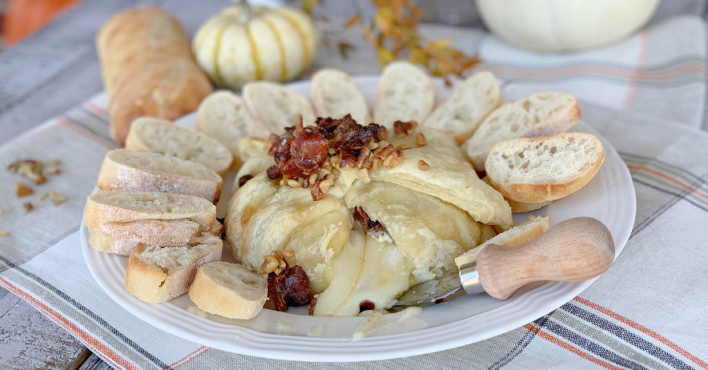 Baked brie with maple syrup in puff pastry on white plate with crispy bacon and walnuts on top and sliced bread around it with the cheese oozing out