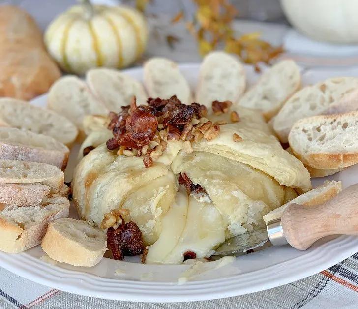 Baked brie with maple syrup in puff pastry on white plate with crispy bacon and walnuts on top and sliced bread around it with the cheese oozing out