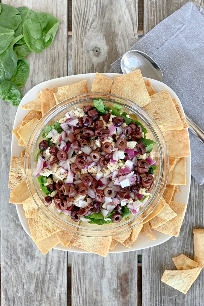 Overhead view of Mediterranean Spread with hummus, spinach, onions and kalamata olives in clear on a white platter with pita chips on a wooden table