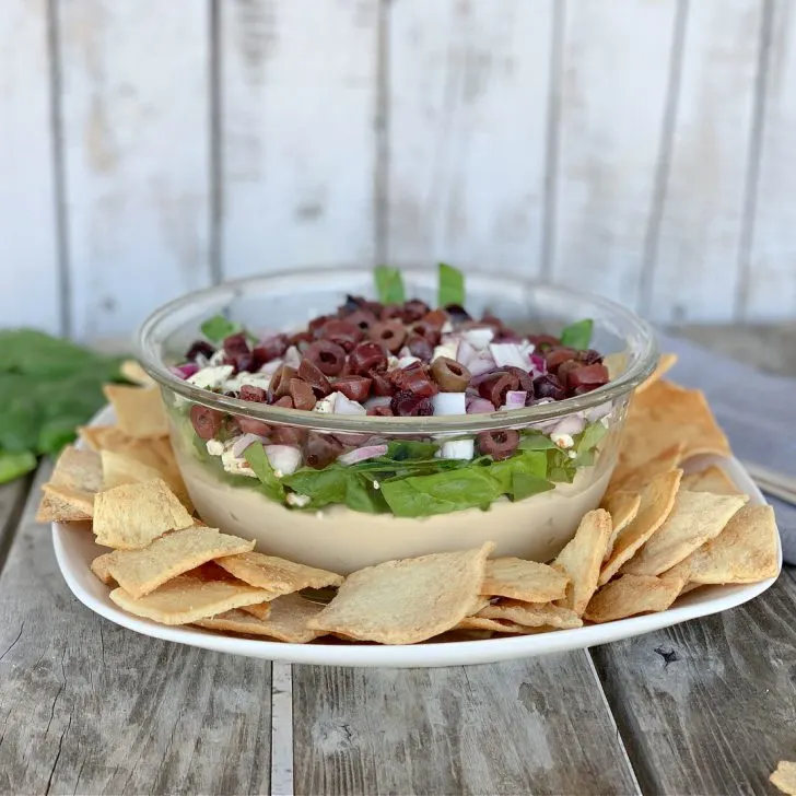 The Mediterranean Spread with hummus, spinach, onions and kalamata olives in clear on a white platter with pita chips on a wooden table