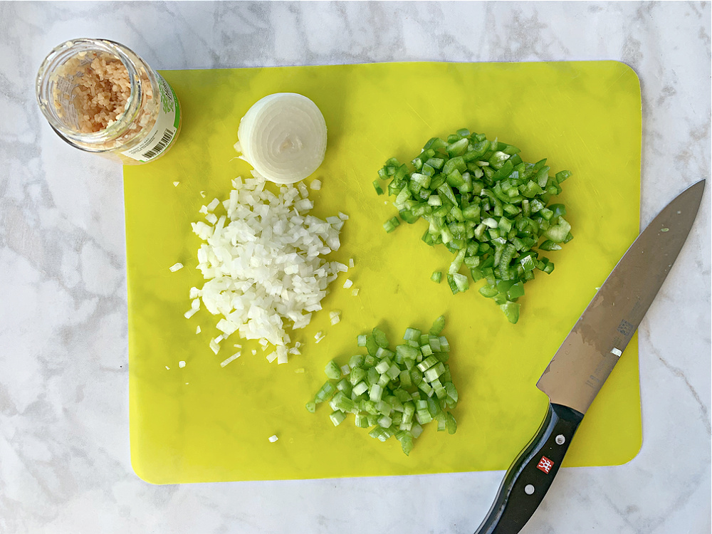 chopped onions, green bell pepper and celery on yellow cutting mat on white countertop