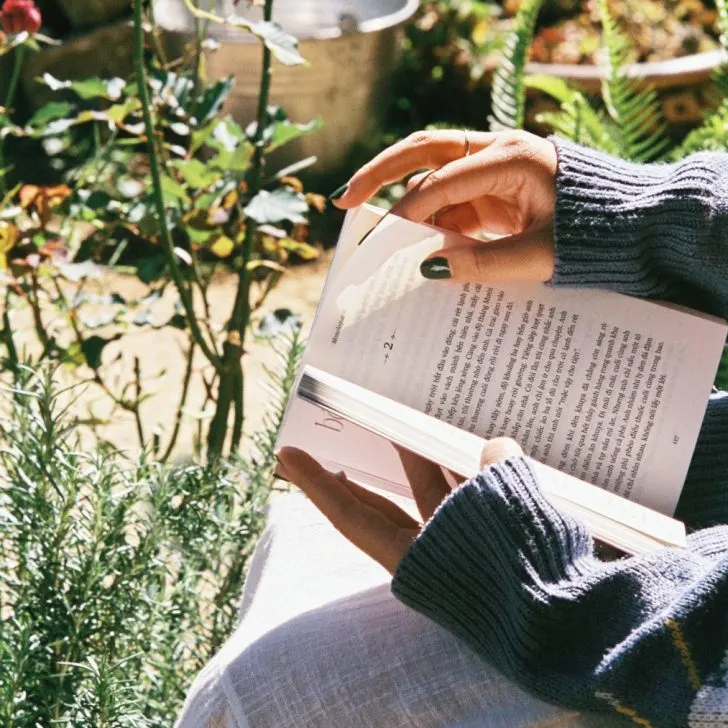 girl reading book outside near green plants